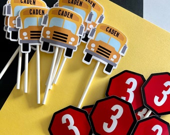 May include: A collection of decorative cupcake toppers featuring yellow school bus illustrations with the name CADEN printed in black, and red octagonal stop signs with the white number 3, all attached to thin white paper sticks.