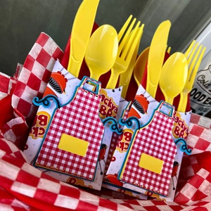 May include: A set of yellow plastic cutlery including knives, spoons, and forks, bundled with paper wraps featuring a red and white checkered apron design, a barbecue grill graphic, and the text Backyard BBQ, all nestled in red and white checkered tissue.