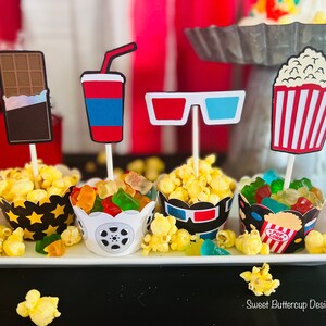 K&ouml;nnte beinhalten: Vier Cupcake-F&ouml;rmchen mit passenden Aufs&auml;tzen im Film-Design, darunter eine Schokoladentafel, ein Getr&auml;nkebecher, eine 3D-Brille und ein Popcorn-Eimer. Die F&ouml;rmchen sind mit Popcorn und Gummib&auml;rchen gef&uuml;llt und stehen auf einem wei&szlig;en Tablett.