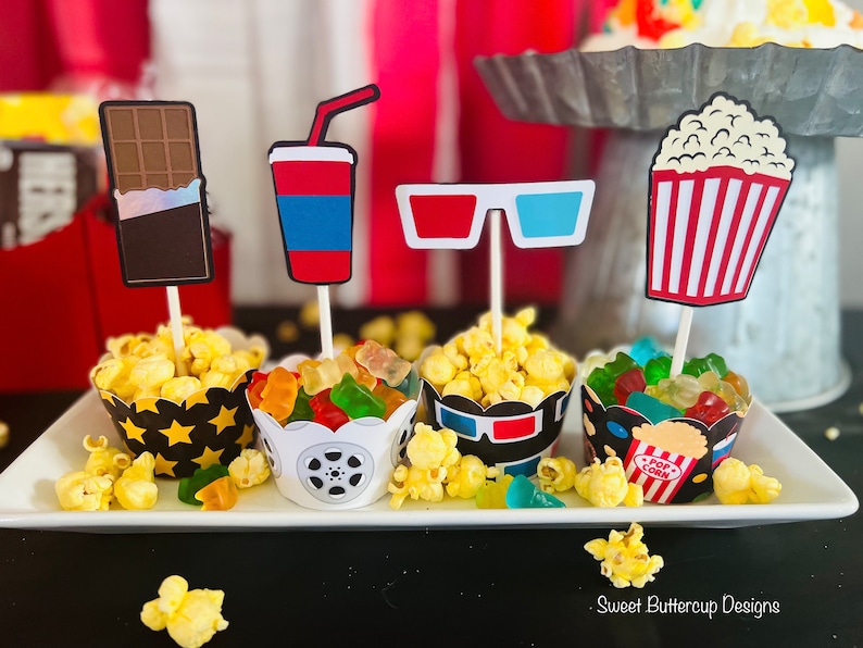 K&ouml;nnte beinhalten: Vier Cupcake-F&ouml;rmchen mit passenden Aufs&auml;tzen im Film-Design, darunter eine Schokoladentafel, ein Getr&auml;nkebecher, eine 3D-Brille und ein Popcorn-Eimer. Die F&ouml;rmchen sind mit Popcorn und Gummib&auml;rchen gef&uuml;llt und stehen auf einem wei&szlig;en Tablett.