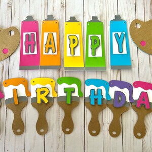 K&ouml;nnte beinhalten: Ein farbenfrohes Geburtstagsbanner mit den Worten "HAPPY BIRTHDAY", geschrieben mit Farbtuben und Pinseln. Das Banner zeigt rosa, orange, gelbe, gr&uuml;ne und blaue Elemente, mit zwei braunen Farbpaletten.