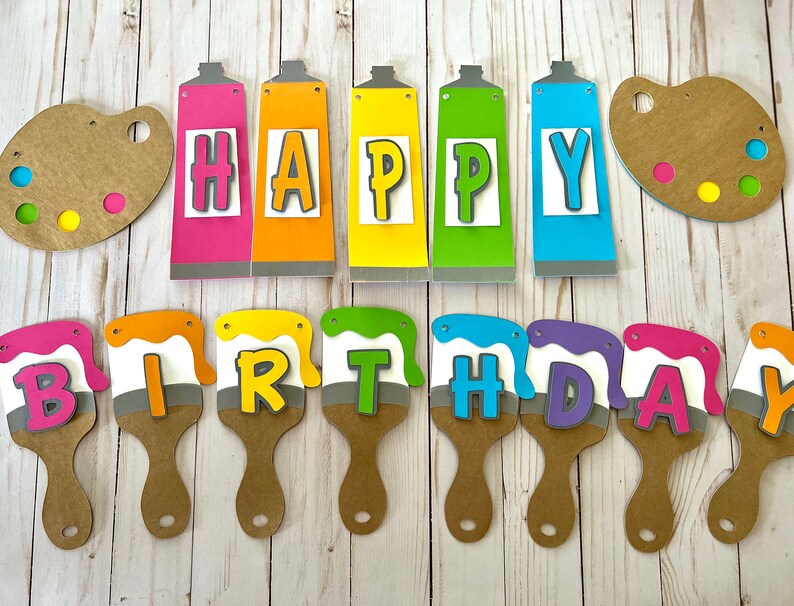 K&ouml;nnte beinhalten: Ein farbenfrohes Geburtstagsbanner mit den Worten "HAPPY BIRTHDAY", geschrieben mit Farbtuben und Pinseln. Das Banner zeigt rosa, orange, gelbe, gr&uuml;ne und blaue Elemente, mit zwei braunen Farbpaletten.