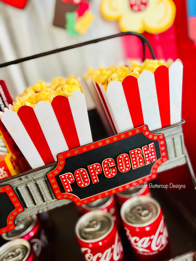 K&ouml;nnte beinhalten: Ein mehrst&ouml;ckiger Metallst&auml;nder mit zwei rot-wei&szlig; gestreiften Popcornbeh&auml;ltern, gef&uuml;llt mit gelbem Popcorn. Ein schwarzes Schild mit rot-wei&szlig; gepunkteten Buchstaben tr&auml;gt die Aufschrift POPCORN. Darunter befinden sich mehrere rote Coca-Cola-Dosen.