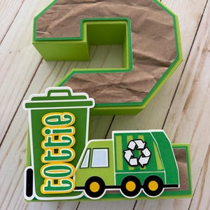 May include: A three-dimensional number two decoration in lime green with a brown, textured paper inlay. It features a green trash bin with the name "Lottie" in yellow and white letters, and a green recycling truck with a white recycling symbol.