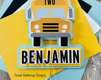 Peut inclure: Une décoration de gâteau en papier superposé représentant un bus scolaire jaune avec le mot TWO sur le toit et une bannière bleu clair affichant le nom BENJAMIN en lettres capitales noires. Le texte Sweet Buttercup Designs est visible.