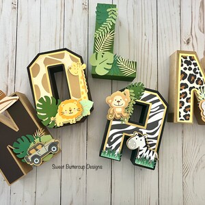 Puede incluir: Letras decorativas marrones, beige y verdes que deletrean el nombre "OLAN". Las letras est&aacute;n decoradas con elementos tem&aacute;ticos de safari, incluyendo un sombrero de safari, jirafa, le&oacute;n, mono, cebra, elefante y jeep. Sobre un fondo de madera clara.
