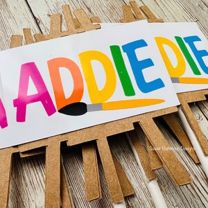 K&ouml;nnte beinhalten: Nahaufnahme mehrerer Partydekorationen zum Thema Kunst. Jedes zeigt eine kleine Staffelei mit einer wei&szlig;en Leinwand mit dem Namen "ADDIE" in bunten Buchstaben. Eine Pinselgrafik ist ebenfalls enthalten. Der Hintergrund ist eine helle Holzoberfl&auml;che.