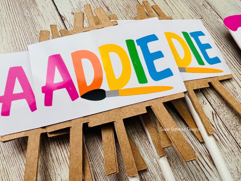 K&ouml;nnte beinhalten: Nahaufnahme mehrerer Partydekorationen zum Thema Kunst. Jedes zeigt eine kleine Staffelei mit einer wei&szlig;en Leinwand mit dem Namen "ADDIE" in bunten Buchstaben. Eine Pinselgrafik ist ebenfalls enthalten. Der Hintergrund ist eine helle Holzoberfl&auml;che.