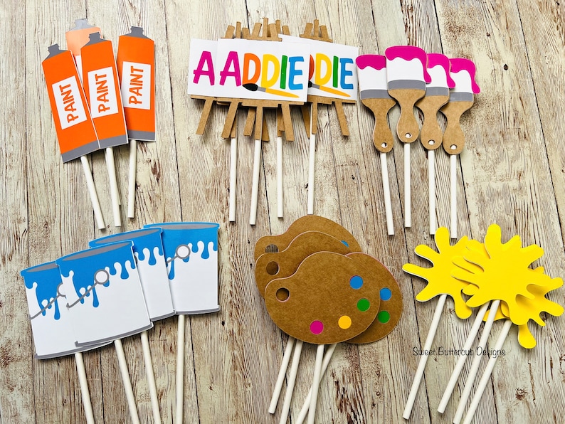 K&ouml;nnte beinhalten: Eine Sammlung von Partydekorationen zum Thema Kunst auf St&auml;ben. Enth&auml;lt orangefarbene Farbtuben, blaue Farbeimer, Staffeleien mit dem Namen "ADDIE", Pinsel mit rosa Farbe, Farbpaletten und gelbe Farbspritzer. Ideal f&uuml;r eine Party zum Thema Kunst.