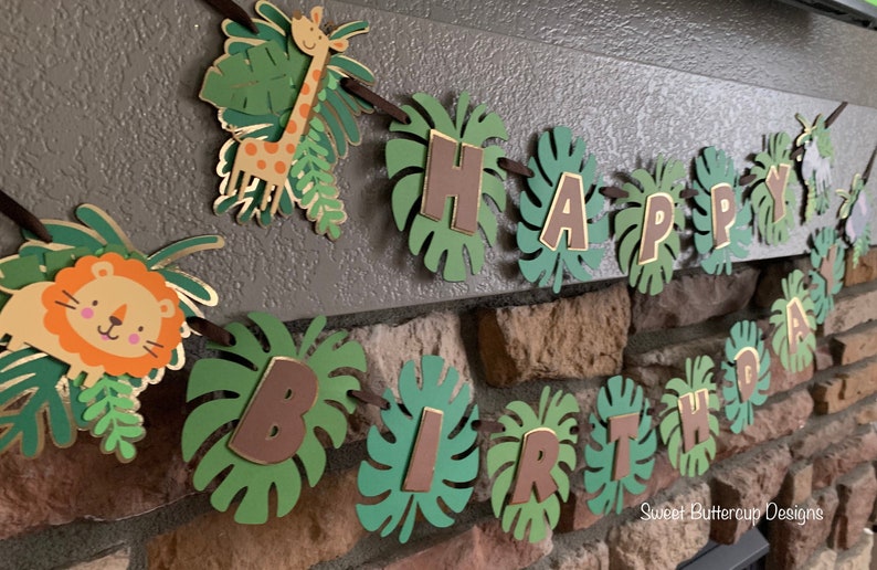 Puede incluir: Una guirnalda de cumplea&ntilde;os con tem&aacute;tica de jungla con las palabras "HAPPY BIRTHDAY" escritas en letras marrones sobre recortes de hojas verdes. La guirnalda incluye una jirafa y un le&oacute;n. La guirnalda est&aacute; ensartada en una cinta marr&oacute;n.