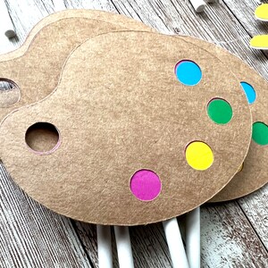 K&ouml;nnte beinhalten: Drei braune K&uuml;nstlerpaletten aus Papier mit farbigen Farbpunkten in Blau, Gr&uuml;n, Gelb und Lila. Jede Palette hat ein Loch f&uuml;r den Daumen und ist an einem wei&szlig;en Stiel befestigt. Die Paletten liegen auf einer Holzoberfl&auml;che.