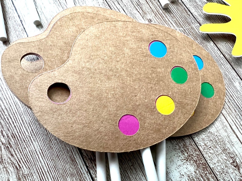 K&ouml;nnte beinhalten: Drei braune K&uuml;nstlerpaletten aus Papier mit farbigen Farbpunkten in Blau, Gr&uuml;n, Gelb und Lila. Jede Palette hat ein Loch f&uuml;r den Daumen und ist an einem wei&szlig;en Stiel befestigt. Die Paletten liegen auf einer Holzoberfl&auml;che.