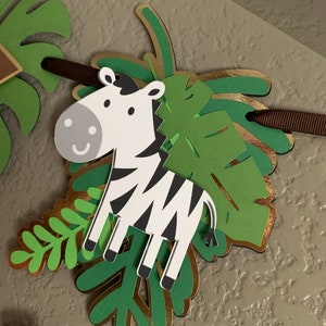 Puede incluir: Un banner decorativo con un cebra de dibujos animados con rayas blancas y negras y una expresi&oacute;n sonriente. La cebra est&aacute; superpuesta a hojas tropicales verdes y doradas, unidas a una cinta marr&oacute;n. El banner tiene un tema de jungla.