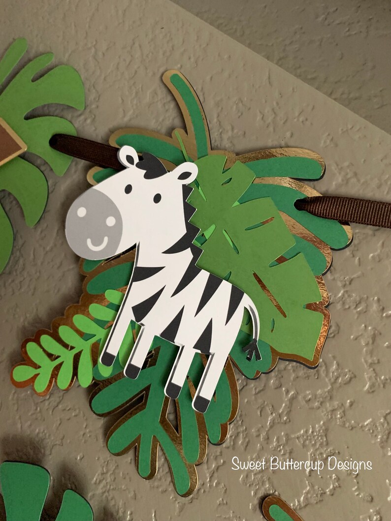 Puede incluir: Un banner decorativo con un cebra de dibujos animados con rayas blancas y negras y una expresi&oacute;n sonriente. La cebra est&aacute; superpuesta a hojas tropicales verdes y doradas, unidas a una cinta marr&oacute;n. El banner tiene un tema de jungla.