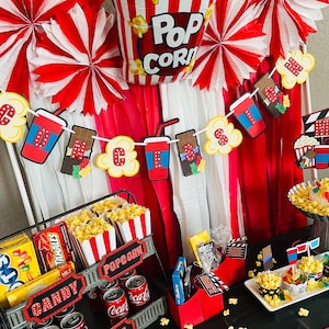 K&ouml;nnte beinhalten: Eine Partydekoration im Kinothema mit einem Popcorn-Ballon, rot-wei&szlig;en Papierf&auml;chern und einer Girlande mit Popcorn-, Soda- und S&uuml;&szlig;igkeiten-Ausschnitten. Ein Metallst&auml;nder enth&auml;lt Popcorn-Boxen und Getr&auml;nkedosen sowie weitere S&uuml;&szlig;igkeiten.