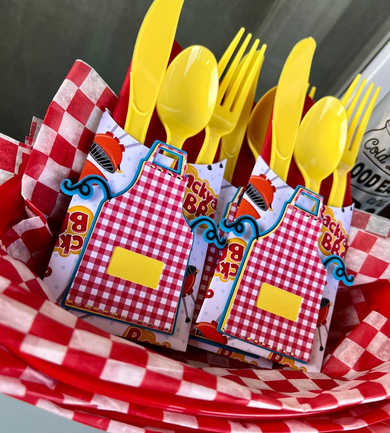 May include: A set of yellow plastic cutlery including knives, spoons, and forks, bundled with paper wraps featuring a red and white checkered apron design, a barbecue grill graphic, and the text Backyard BBQ, all nestled in red and white checkered tissue.