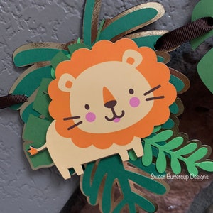 Puede incluir: Un cartel decorativo con un le&oacute;n de dibujos animados, con una melena naranja, mejillas rosas y una expresi&oacute;n amigable. El le&oacute;n est&aacute; rodeado de hojas de papel verdes y doradas. Se adjunta una cinta marr&oacute;n para colgarlo. El cartel tiene un tema de jungla o safari.