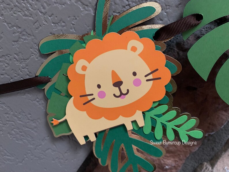 Puede incluir: Un cartel decorativo con un le&oacute;n de dibujos animados, con una melena naranja, mejillas rosas y una expresi&oacute;n amigable. El le&oacute;n est&aacute; rodeado de hojas de papel verdes y doradas. Se adjunta una cinta marr&oacute;n para colgarlo. El cartel tiene un tema de jungla o safari.