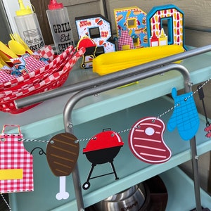 May include: A light blue metal cart holding a red plastic basket with yellow cutlery, two clear squeeze bottles, and decorative letters spelling BBQ. A hanging banner features paper cutouts of an apron, meat, a grill, a steak, a mitt, and condiments.