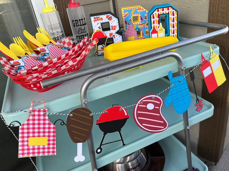 May include: A light blue metal cart holding a red plastic basket with yellow cutlery, two clear squeeze bottles, and decorative letters spelling BBQ. A hanging banner features paper cutouts of an apron, meat, a grill, a steak, a mitt, and condiments.