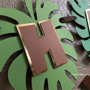 Puede incluir: Un cartel hecho a mano con las letras "HA" y una letra marr&oacute;n "H" con ribete dorado. Las letras est&aacute;n sobre recortes de hojas de monstera verdes. El cartel est&aacute; ensartado con cinta marr&oacute;n. El texto "Sweet Buttercup Designs" es visible.