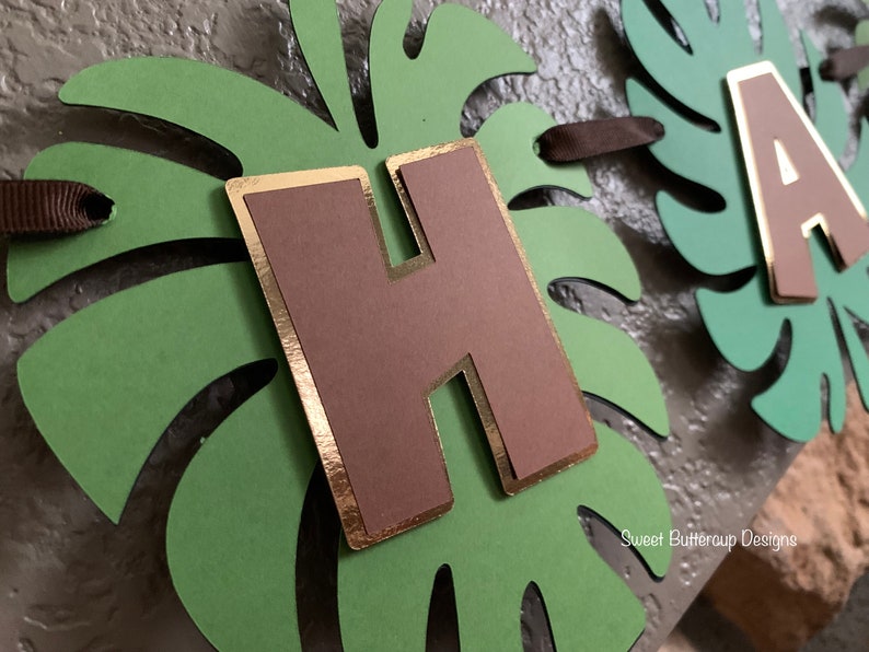 Puede incluir: Un cartel hecho a mano con las letras "HA" y una letra marr&oacute;n "H" con ribete dorado. Las letras est&aacute;n sobre recortes de hojas de monstera verdes. El cartel est&aacute; ensartado con cinta marr&oacute;n. El texto "Sweet Buttercup Designs" es visible.