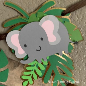 Puede incluir: Un bander&iacute;n de elefante gris con orejas rosas y una cara sonriente, rodeado de hojas verdes y doradas. El bander&iacute;n est&aacute; ensartado en una cinta marr&oacute;n. El texto "Sweet Buttercup Designs" es visible.