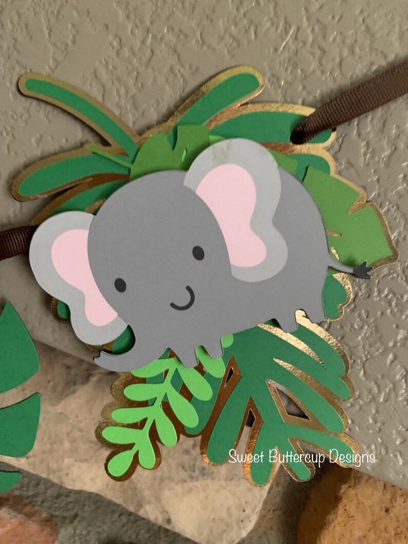 Puede incluir: Un bander&iacute;n de elefante gris con orejas rosas y una cara sonriente, rodeado de hojas verdes y doradas. El bander&iacute;n est&aacute; ensartado en una cinta marr&oacute;n. El texto "Sweet Buttercup Designs" es visible.