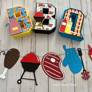 May include: Three-dimensional letters spelling BBQ, decorated with food patterns, alongside a hanging banner featuring cutouts of a red and white checkered apron, a chicken leg, a charcoal grill, a steak, a blue oven mitt, and condiment bottles.