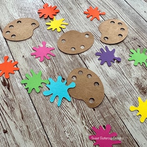 K&ouml;nnte beinhalten: Bastelbedarf, darunter braune Holzmalerpaletten und farbenfrohe Papierspritzer in Orange, Gelb, Rosa, Gr&uuml;n, Blau und Lila. Die Artikel sind auf einer rustikalen, hellen Holzoberfl&auml;che angeordnet.