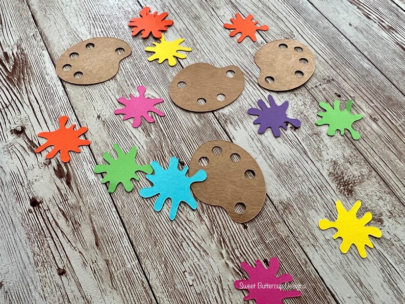 K&ouml;nnte beinhalten: Bastelbedarf, darunter braune Holzmalerpaletten und farbenfrohe Papierspritzer in Orange, Gelb, Rosa, Gr&uuml;n, Blau und Lila. Die Artikel sind auf einer rustikalen, hellen Holzoberfl&auml;che angeordnet.