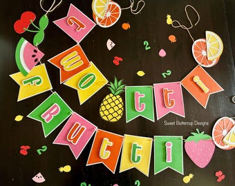 May include: A decorative paper banner featuring the text "TWOOTTI FRUTTI" on colorful pennants interspersed with paper cutouts of a pineapple, strawberry, watermelon, cherries, and citrus slices, with the text "Sweet Buttercup Designs" visible.