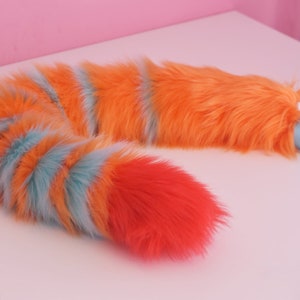 May include: A fluffy orange, blue, and red striped tail with a blue ribbon attached. The tail is made of faux fur and is designed to be worn as a costume accessory.