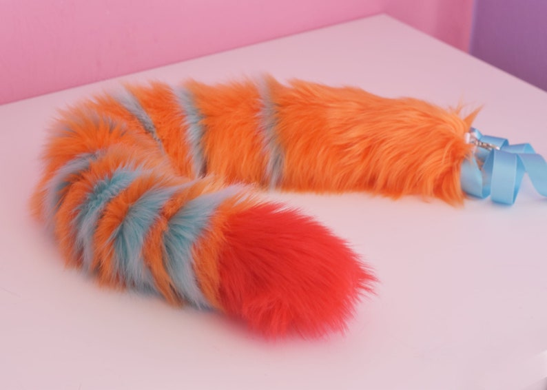 May include: A fluffy orange, blue, and red striped tail with a blue ribbon attached. The tail is made of faux fur and is designed to be worn as a costume accessory.