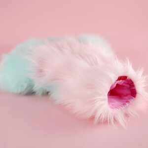 May include: A fluffy, pastel pink and blue faux fur cat bed with a pink lining. The bed is shaped like a tunnel and is perfect for cats to curl up in.