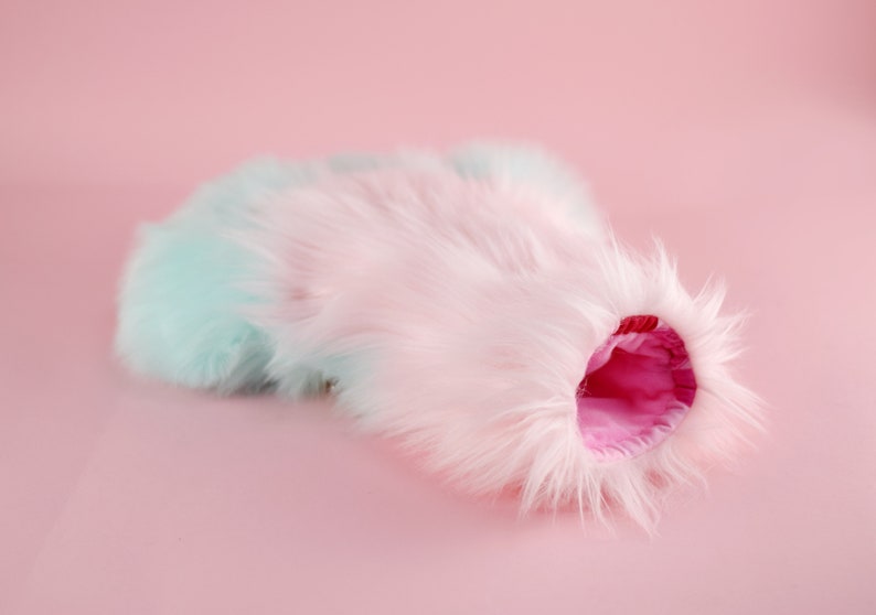 May include: A fluffy, pastel pink and blue faux fur cat bed with a pink lining. The bed is shaped like a tunnel and is perfect for cats to curl up in.