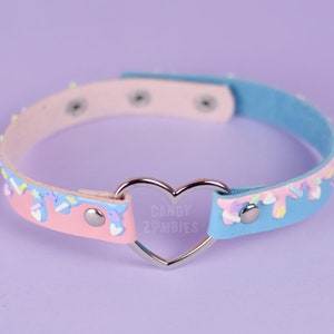 May include: A pastel choker necklace with a heart-shaped centre. The necklace has pink and blue sections with decorative icing details. The text "CANDY ZOMBIES" is visible on the heart. The necklace has silver-coloured hardware.