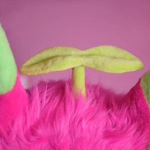 May include: A pink furry plush toy with a green leaf-shaped headpiece. The headpiece is attached to the toy's head with a green stem.