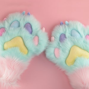 May include: A pair of light blue fuzzy paw-shaped hand warmers with pink and yellow accents. The hand warmers have a fluffy white cuff.