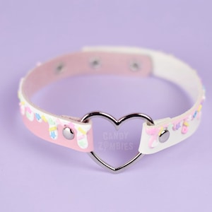 May include: A pastel pink and white choker with a silver heart-shaped center. The choker features a decorative design with sprinkles and the text "CANDY ZOMBIES".