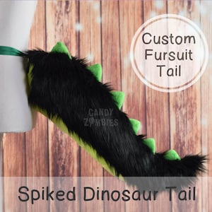 Könnte beinhalten: Schwarzer und grüner, maßgeschneiderter Fellschwanz mit Stacheln. Der Schwanz besteht aus schwarzem Fell mit grünen Stacheln und einer grün-gelben Basis. Der Text "Custom Fursuit Tail" ist in Weiß auf einem schwarzen Kreis geschrieben. Der Text "Spiked Dinosaur Tail" ist in Weiß auf schwarzem Hintergrund geschrieben.