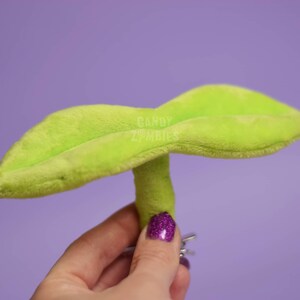 May include: A green plush toy shaped like a plant with two large leaves and a stem. The toy is held in a hand with purple nail polish.