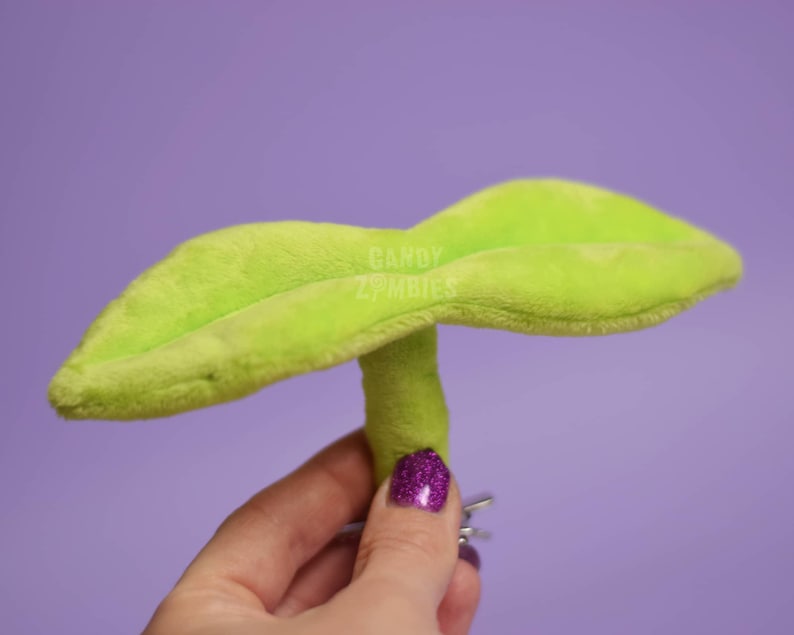 May include: A green plush toy shaped like a plant with two large leaves and a stem. The toy is held in a hand with purple nail polish.