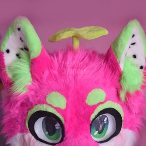 May include: Pink and green furry plush toy with a watermelon pattern on the ears and a green leaf on top of the head. The toy has large green eyes and a white muzzle.