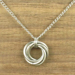 May include: A silver-toned necklace with a pendant featuring a series of interlocking rings. The chain is a delicate, linked design. The pendant's rings are smooth and polished, creating a modern and minimalist aesthetic. The necklace is displayed against a wooden background.