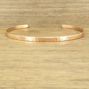 May include: A hammered copper cuff bracelet. The open-ended bracelet has a textured surface and a warm, metallic color. The bracelet is approximately 0.25 inches wide.