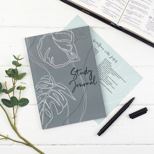 Puede incluir: Un diario de estudio gris con un diseño floral blanco y el texto "Study Journal" en la portada. El diario está sobre una superficie de madera blanca con un bolígrafo negro y una planta verde.