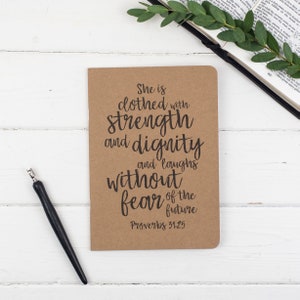 May include: A brown notebook with the quote "She is clothed with strength and dignity and laughs without fear of the future" and the scripture reference "Proverbs 31:25" printed in black.