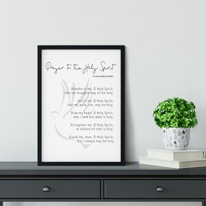 May include: A framed print featuring the prayer "Prayer to the Holy Spirit" by St. Augustine of Hippo. The black frame encloses a white background with the prayer in black text and a dove illustration. A small potted plant and books are on a dark grey table.