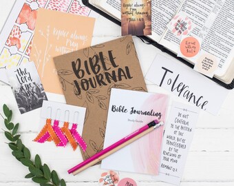 Può includere: Un diario marrone con le parole "Bible Journal" scritte in bianco sulla copertina. Il diario è circondato da vari oggetti, tra cui una Bibbia, una guida allo studio, una matita e diversi ritagli di carta colorati con citazioni ispiratrici.